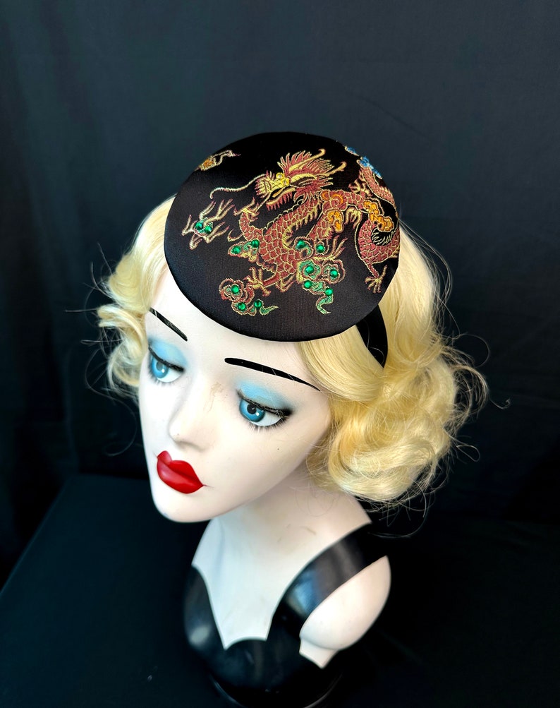 May include: A black satin hat featuring a gold and red embroidered dragon design. The dragon has green accents and is surrounded by gold and green embellishments.