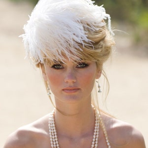 May include: A woman wearing a white feather hat and a pearl necklace. She has blonde hair and is looking at the camera.