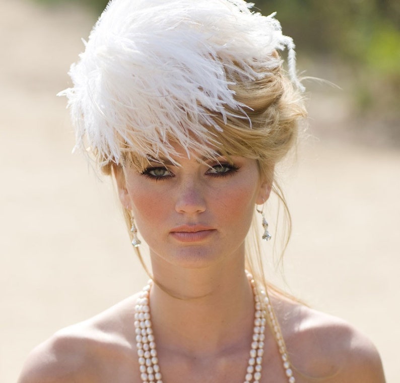 May include: A woman wearing a white feather hat and a pearl necklace. She has blonde hair and is looking at the camera.