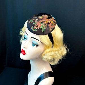 May include: A black satin hat with a red and gold embroidered dragon design. The dragon has green jewels on its body.