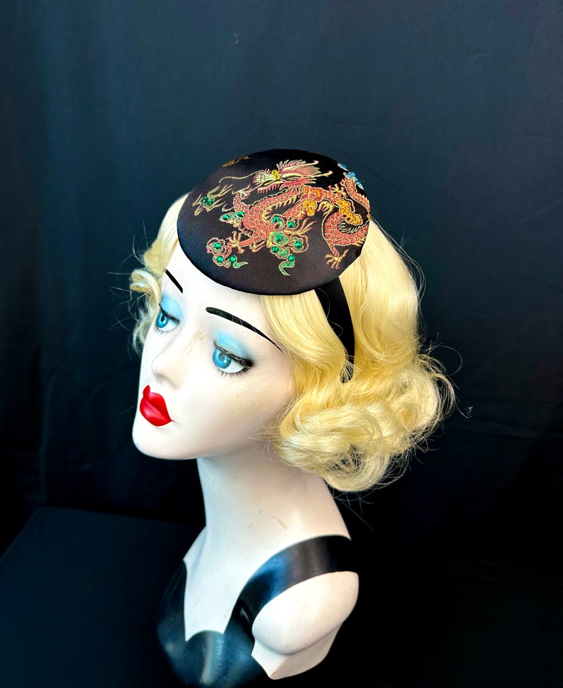 May include: A black satin hat with a red and gold embroidered dragon design. The dragon has green jewels on its body.