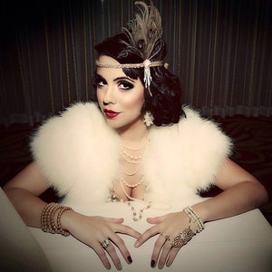 May include: A woman wearing a white fur stole and a beaded headband with a feather accent. She is wearing a pearl necklace and a gold bracelet. Her nails are painted with a black and white design.