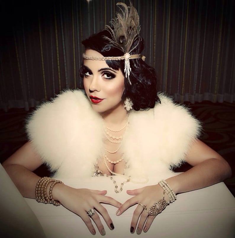 May include: A woman wearing a white fur stole and a beaded headband with a feather accent. She is wearing a pearl necklace and a gold bracelet. Her nails are painted with a black and white design.