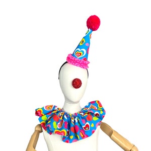 May include: A blue and pink clown hat with a red pom-pom on top. The hat has a pink ruffle around the base and a pattern of colorful hearts. A red clown nose is attached to a white mannequin head wearing a blue and pink ruffled collar with a heart pattern.