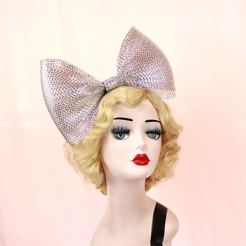 May include: A large silver sparkly bow headband. The bow is made of a glittery material and is tied in a large, dramatic knot. The headband is worn on a mannequin with blonde hair.