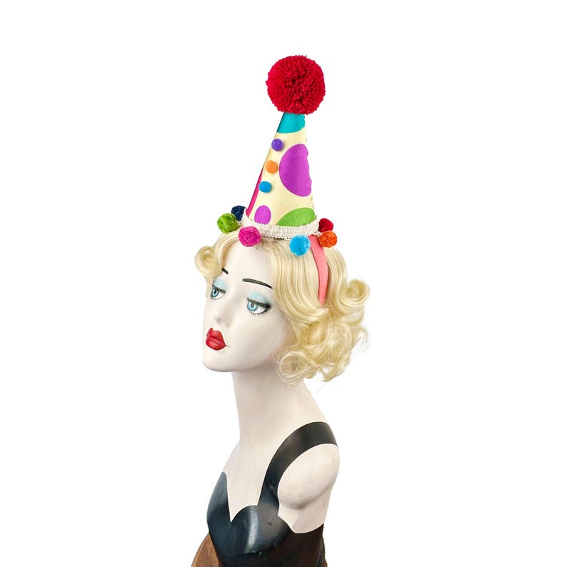 Pode incluir: Um chap&eacute;u de festa colorido com um pompom vermelho no topo. O chap&eacute;u &eacute; decorado com bolinhas de v&aacute;rias cores, incluindo rosa, azul, verde e amarelo. O chap&eacute;u &eacute; usado por um manequim com cabelo loiro encaracolado.