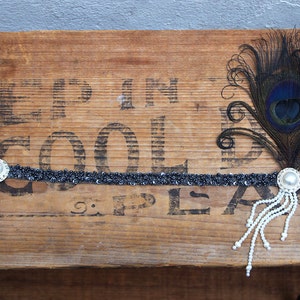 May include: A black headband with a beaded design and a pearl accent. A black feather with a peacock eye design is attached to the headband.