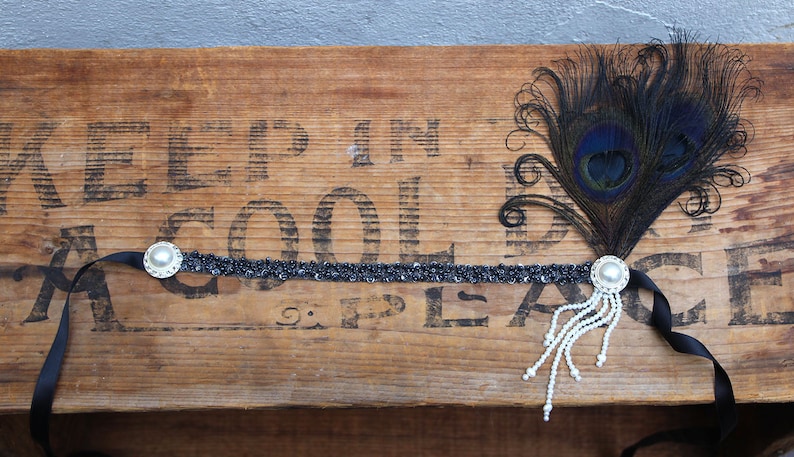 May include: A black headband with a beaded design and a pearl accent. A black feather with a peacock eye design is attached to the headband.