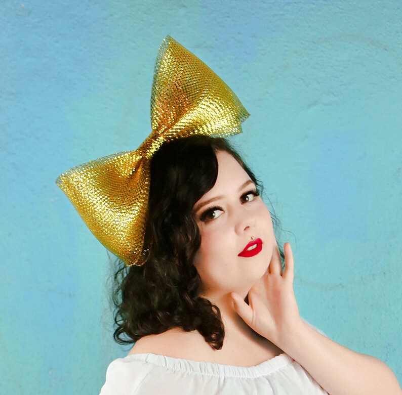 May include: A woman with dark curly hair is wearing a large gold bow on her head. She is wearing a white off-the-shoulder top and has red lipstick on.