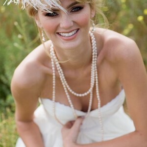 May include: A bride wearing a white strapless dress with a pearl necklace and a white feather headpiece. She is smiling and looking at the camera.