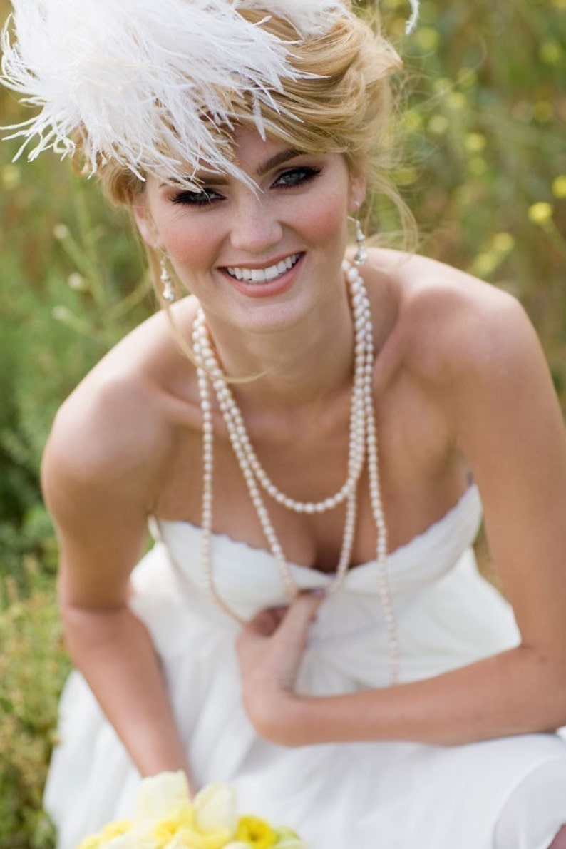 May include: A bride wearing a white strapless dress with a pearl necklace and a white feather headpiece. She is smiling and looking at the camera.