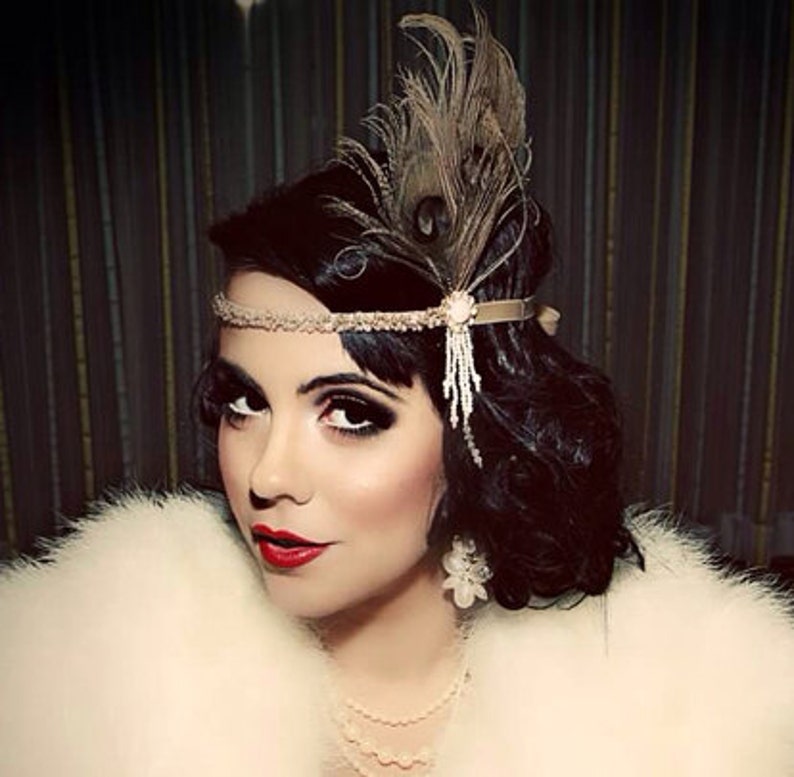 May include: A woman with dark hair styled in a 1920s bob wears a headband with a feather and beaded fringe. She is wearing a white fur stole and a pearl necklace.