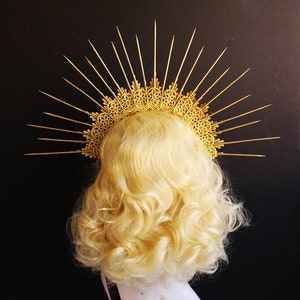 May include: A gold halo headpiece with a delicate, intricate design and sharp, pointed rays. The halo is worn on a blonde wig.