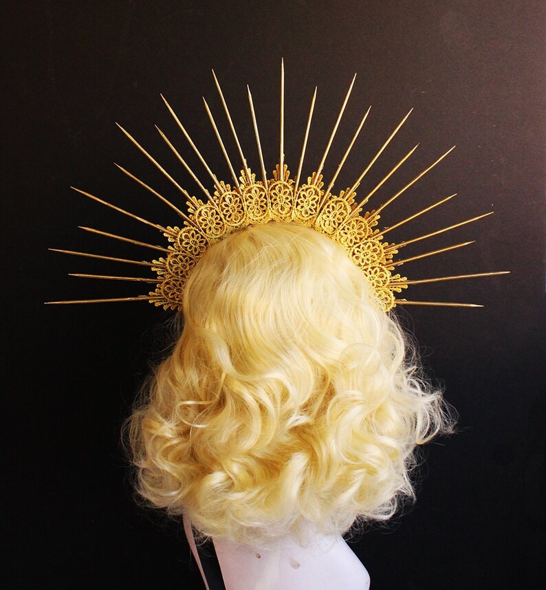 May include: A gold halo headpiece with a delicate, intricate design and sharp, pointed rays. The halo is worn on a blonde wig.
