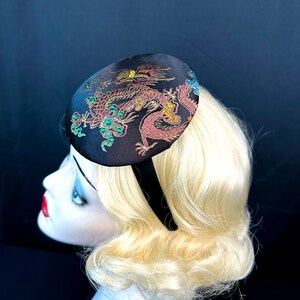 May include: A black satin headband with a round, embroidered fabric topper featuring a gold and red dragon design with green accents.
