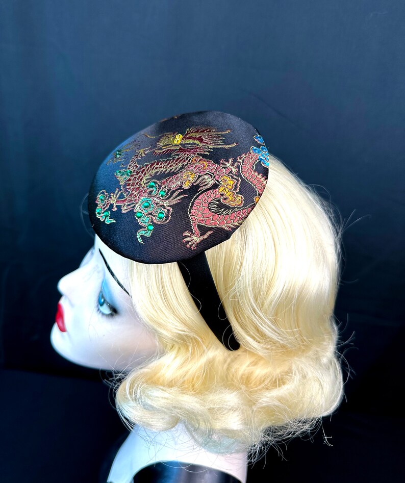 May include: A black satin headband with a round, embroidered fabric topper featuring a gold and red dragon design with green accents.