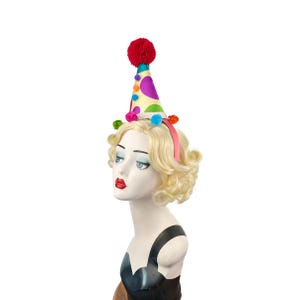 Pode incluir: Uma faixa de cabelo com um chap&eacute;u de palha&ccedil;o colorido com um pompom vermelho no topo. O chap&eacute;u &eacute; decorado com bolinhas coloridas e pompons.