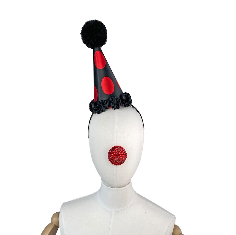 May include: A black and red clown hat with a black pom-pom on top. The hat has red polka dots and a black ruffle around the base. A red jeweled clown nose is attached to a white mannequin head.