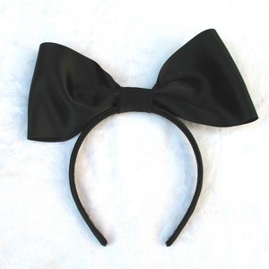 May include: A black satin bow headband. The headband is made of a thin black elastic band and has a large black satin bow attached to it.