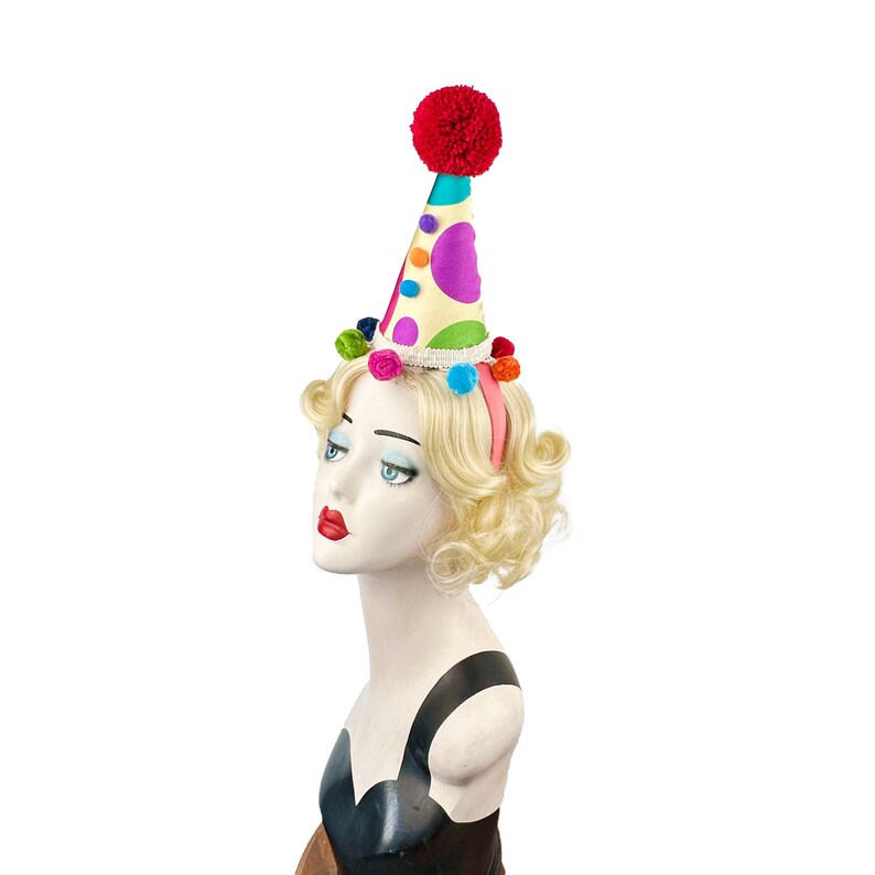 Pode incluir: Um chap&eacute;u de festa colorido com um pompom vermelho no topo. O chap&eacute;u &eacute; decorado com bolinhas brancas e pompons multicoloridos ao redor da base.