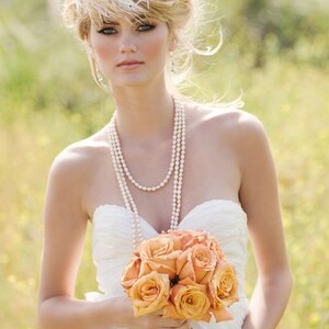 May include: A bride in a white strapless wedding dress holds a bouquet of peach-colored roses. She is wearing a pearl necklace and a white feather headpiece.
