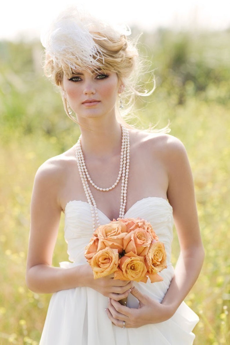 May include: A bride in a white strapless wedding dress holds a bouquet of peach-colored roses. She is wearing a pearl necklace and a white feather headpiece.