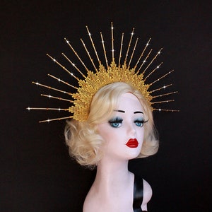 May include: A gold crown with a sunburst design and sparkling rhinestones. The crown is worn on a mannequin with blonde hair.