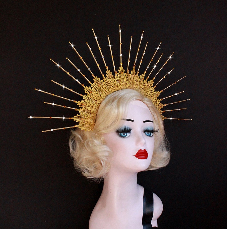 May include: A gold crown with a sunburst design and sparkling rhinestones. The crown is worn on a mannequin with blonde hair.