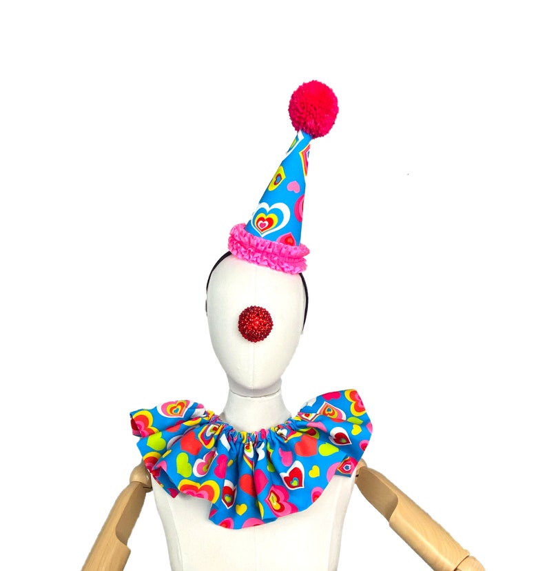 May include: A blue and pink clown costume with a heart pattern. The costume includes a hat with a pink pom-pom, a red clown nose, and a ruffled collar.