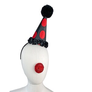 May include: A black and red polka dot party hat with a black pom pom on top. The hat is on a white mannequin head with a red, sparkly clown nose.