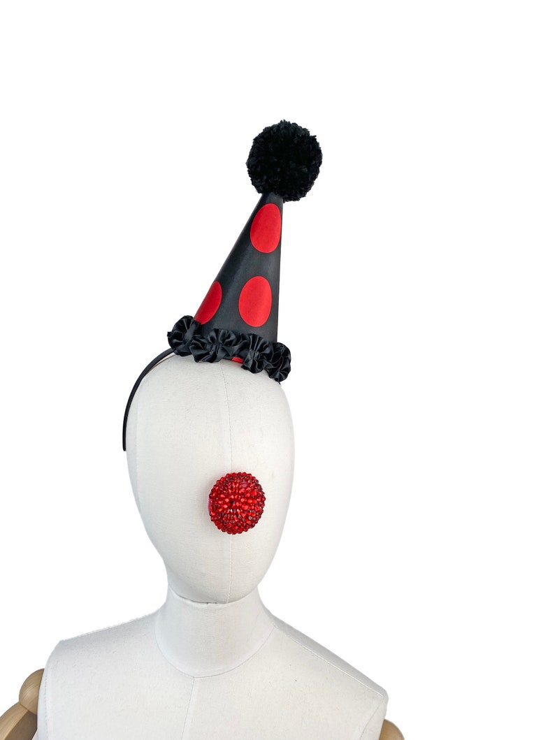 May include: A black and red polka dot party hat with a black pom pom on top. The hat is on a white mannequin head with a red, sparkly clown nose.