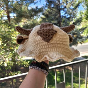 Pode incluir: Um chapéu de crochê em forma de bisão, com base creme e detalhes marrons para os chifres e marcas. O chapéu tem uma aba com babados. O fundo mostra árvores verdes e céu azul.
