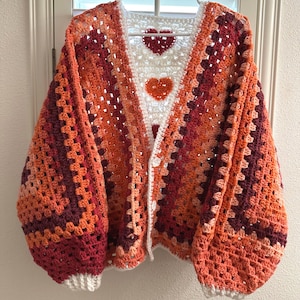 May include: A handmade crochet cardigan in shades of orange, burgundy, and white. The open-front sweater features a granny square design with heart-shaped accents on the front. The sleeves are long and voluminous, and the cardigan is hung on a white hanger.