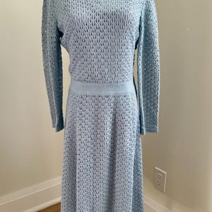May include: A pale blue knitted dress with a collared neckline and long sleeves. The dress has a textured, open-weave pattern and a fitted waist. The skirt is a midi length. The dress is displayed on a mannequin.