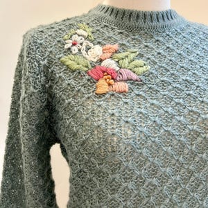 May include: A light blue sweater with a textured knit pattern and long sleeves. The sweater features a floral embroidery design on the upper left chest, with pink, white, and orange flowers and green leaves.