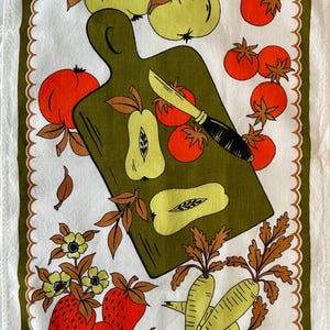 May include: A white kitchen towel with a scalloped olive green border featuring a central olive green cutting board, a knife, and various illustrations of red tomatoes, light green apples, strawberries, and pale yellow root vegetables.