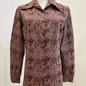 May include: A long-sleeved button-down shirt with a large collar. The shirt features a repeating pattern of floral and geometric shapes in shades of brown, tan, and burgundy. The shirt is displayed on a mannequin.