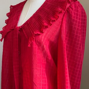 May include: A bright red blouse with a large, ruffled collar and long sleeves. The fabric has a subtle checkered pattern. The blouse has a V-neckline and buttons down the front.