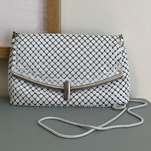 May include: A white mesh handbag featuring a rectangular shape with a metallic silver-toned curved bar closure at the front. The bag is constructed from small, interlocking square tiles and includes a thin, flexible silver-toned shoulder strap.