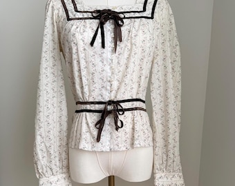 Könnte beinhalten: Eine weiße Bluse mit floralem Muster, langen Ärmeln, eckigem Ausschnitt und braunem Samtbesatz. Die Bluse hat eine braune Samtschleife am Ausschnitt und in der Taille und wird auf einer Schaufensterpuppe präsentiert.