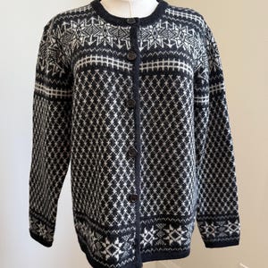 Könnte beinhalten: Eine schwarz-weiß gemusterte Strickjacke mit Knopfleiste. Der Pullover hat ein Rauten- und geometrisches Design, einen runden Ausschnitt und lange Ärmel. Der Pullover wird auf einer Schaufensterpuppe präsentiert.