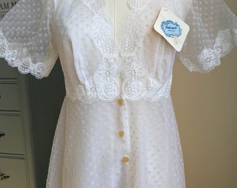Könnte beinhalten: Ein weißes, transparentes Kleid mit Swiss-Dot-Muster, kurzen Ärmeln, einem tiefen V-Ausschnitt mit aufwendiger floraler Spitzenborte und drei kleinen Knöpfen auf der Vorderseite. Ein weißes Etikett mit dem Text French Maid ist am Ausschnitt befestigt.