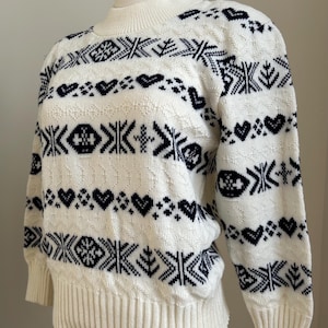 May include: Cream-colored sweater with a mock neck and long sleeves. The sweater features a repeating pattern of navy blue geometric shapes, hearts, and snowflakes. The cuffs and hem are ribbed.