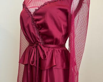 Könnte beinhalten: Burgunderfarbenes Vintage-Kleid auf einer Schaufensterpuppe. Das Kleid hat einen hohen Rüschenkragen, transparente gepunktete Ärmel und ein Wickeloberteil mit Bindeband. Der Rock hat ein geschichtetes Schößchen-Design und schafft eine klassische Silhouette.