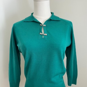 Puede incluir: Jersey verde turquesa con cuello y mangas tres cuartos. El jersey presenta un detalle de ojal en el escote, asegurado por dos cierres rectangulares plateados. La prenda se exhibe sobre un maniquí blanco.
