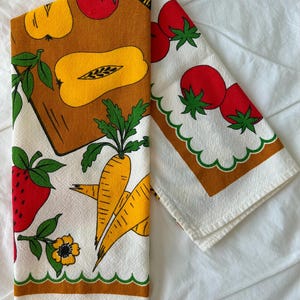 May include: A white kitchen towel featuring a colorful print of red strawberries, yellow squash, orange carrots, and red tomatoes. The design includes green leaves and scalloped green borders with brown rectangular accents on a textured fabric.