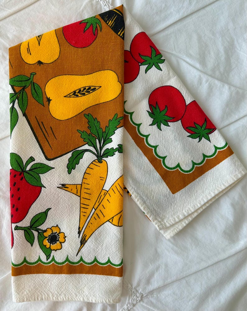 May include: A white kitchen towel featuring a colorful print of red strawberries, yellow squash, orange carrots, and red tomatoes. The design includes green leaves and scalloped green borders with brown rectangular accents on a textured fabric.