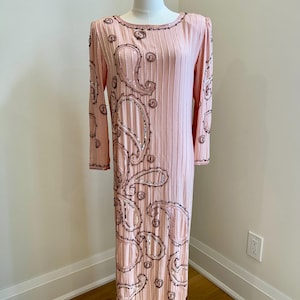 Puede incluir: Vestido rosa pálido de manga larga, cuello redondo y largo midi. El vestido presenta pliegues verticales y un diseño de cachemira adornado con lentejuelas y ribetes marrón oscuro. El vestido se exhibe en un maniquí.