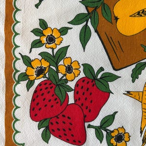 May include: A vintage white kitchen towel with a repeating pattern of red strawberries, yellow flowers, and green leaves. The towel has a brown border with a scalloped edge. The design also includes yellow apples.