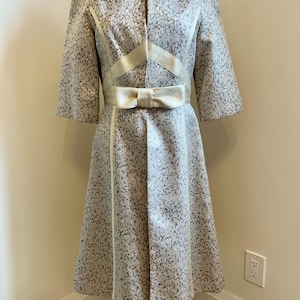 May include: A white and gray patterned dress with a gold bow belt. The dress has a fitted bodice and a flared skirt. The sleeves are three-quarter length.